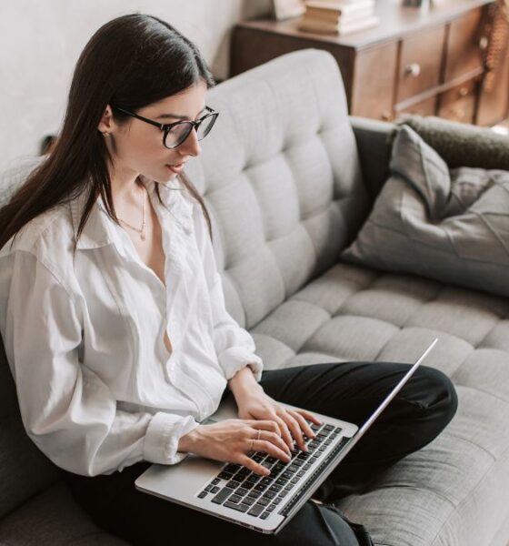 A woman with glasses sitting cross legged on a gray couch working on laptop