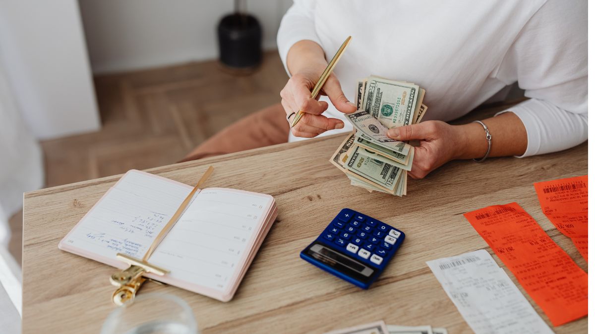 Closeup of a women's hands counting money next to a calculator, receipts, and open notebook